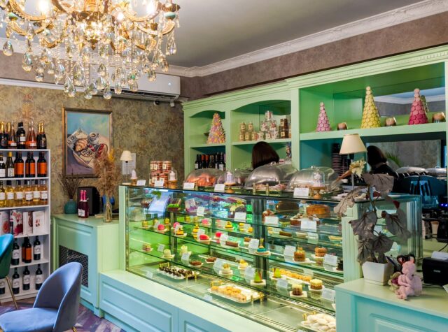 Elegant cafe interior with chandelier, mint-green shelves filled with pastries, wine bottles, and decorative items in Tbilisi.