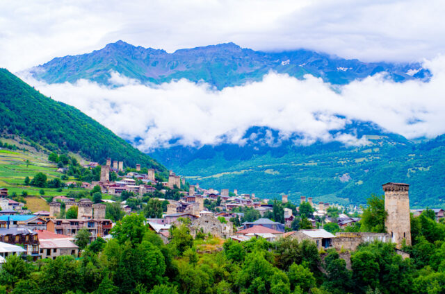 Scenic view of Mestia in Svaneti, Georgia, with stone medieval Svan towers rising among traditional houses, surrounded by lush green hills and dramatic mountains partly covered by clouds.