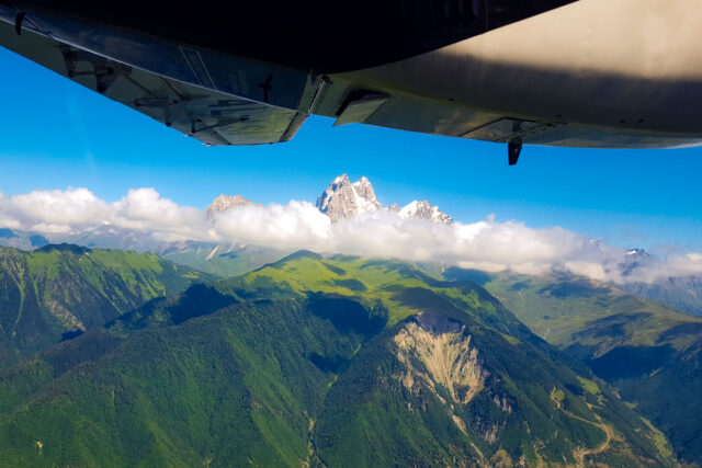 Aerial view of Mount Ushba in the Caucasus Mountains, Svaneti, Georgia, captured from a small plane. The snow-capped twin peaks rise dramatically above a layer of clouds, with rolling green hills and deep forested valleys spread below under a clear blue sky.