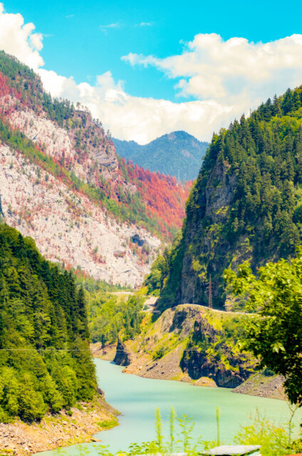 Turquoise river winding through a steep gorge surrounded by forested cliffs and rocky slopes with patches of red trees, on the scenic mountain road to Mestia in Svaneti, Georgia. Photo taken from the minibus