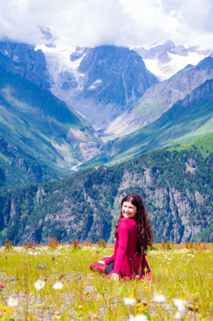 Smiling woman with long dark hair wearing a dark red jacket sits in a wildflower meadow on Mount Tetnuldi in Svaneti, Georgia, with rugged green valleys and snow-capped mountains in the background.