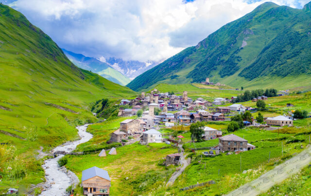 Scenic view of Ushguli village in Svaneti, Georgia, surrounded by lush green valleys and towering mountains. The medieval stone defensive towers and traditional stone houses Svaneti towers are scattered along the hillside, with a river winding through the foreground under a dramatic cloudy sky.