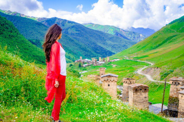 Woman with long dark hair in a red shawl and pants overlooking the medieval stone towers of Ushguli, Svaneti, with lush green mountains and a winding road in the backdrop,