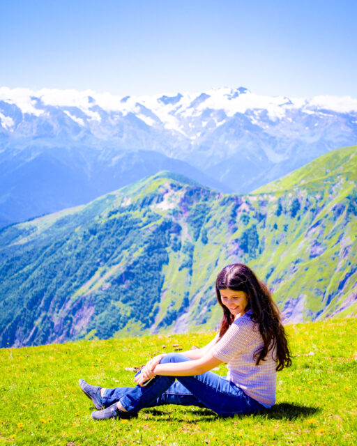 Woman with long dark hair wearing a creamy colored short-sleeve top and blue jeans sitting on a grassy alpine meadow, smiling against a backdrop of green ridges and the snow-capped Caucasus Mountains in Svaneti, Georgia.