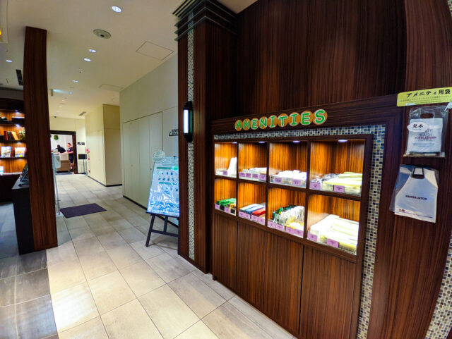 Amenities corner at Hotel Keihan in Osaka with a wooden display cabinet offering free guest items like toothbrushes, combs, razors, and tea bags under a brightly lit “AMENITIES” sign.