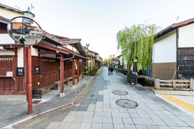 Scenic street in Hida-Furukawa, Japan, featuring traditional wooden townhouses, a willow tree above a narrow canal, and tiled stone pavement with manhole covers. Two people walk in the distance, while a convex mirror reflects nearby buildings. On the left side, wooden posts with benches display toilet signs for public restrooms.