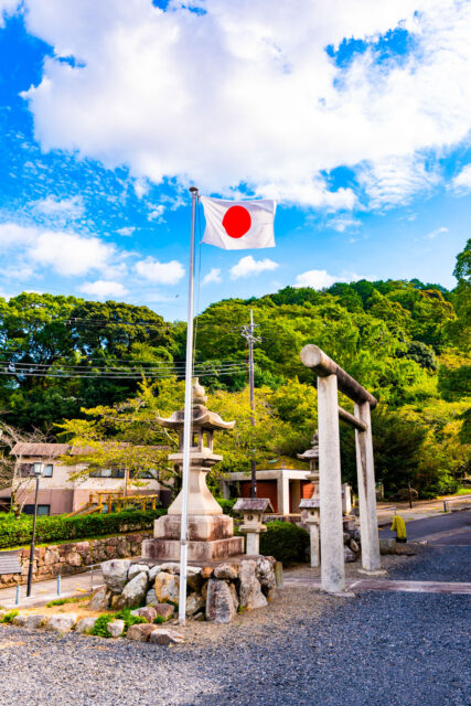 Japanese flag flying on a tall pole next to a stone lantern and torii gate at a Shinto shrine in Ōtsu, Japan, with green forested hills and a partly cloudy blue sky in the background.