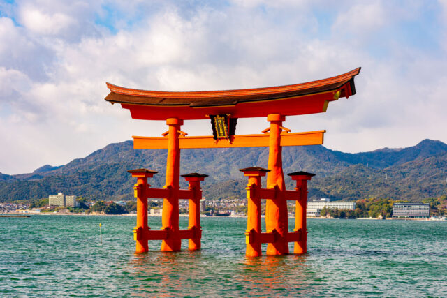 The iconic vermilion Great Torii Gate of Itsukushima Shrine standing in the sea off Miyajima Island, Hiroshima, Japan. The gate rises from the water against a backdrop of forested mountains and a partly cloudy sky, with buildings along the shoreline visible in the distance.