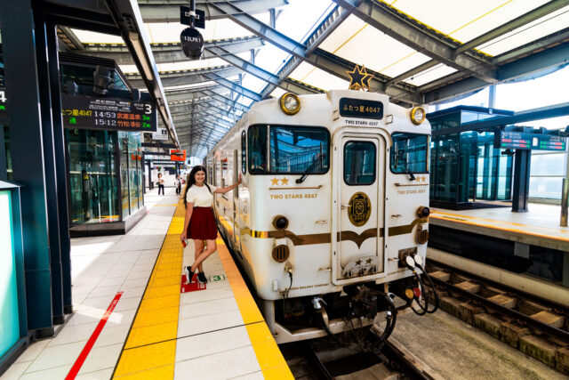 White and gold Two Stars 4047 sightseeing train at Saga Station in Japan with a woman standing beside it on the platform