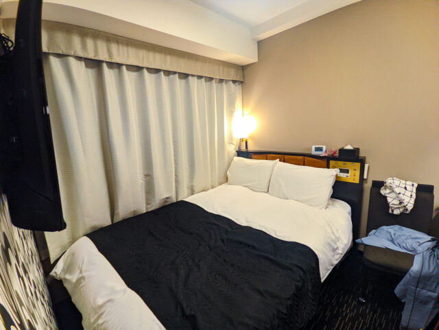 Compact double room at APA Hotel Hakata in Japan, a typical small-sized Japanese business hotel room with a neatly made bed, black duvet, white pillows, and soft warm lighting.