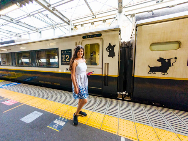 A woman with long hair in a white dress with blue floral patterns standing on the platform next to the Aso Boy! sightseeing train in Kyushu, Japan - white exterior decorated with Kuro-chan dog mascot illustrations on the cars.