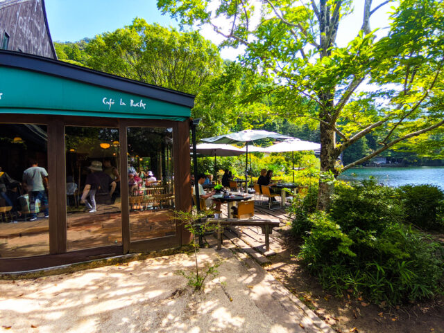 Exterior of Café La Ruche in Yufuin, Oita, Japan with outdoor seating under umbrellas overlooking a Kinrin lakeside surrounded by greenery.