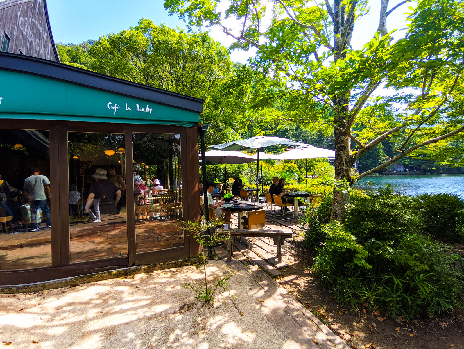 Exterior of Café La Ruche in Yufuin, Oita, Japan with outdoor seating under umbrellas overlooking a Kinrin lakeside surrounded by greenery.