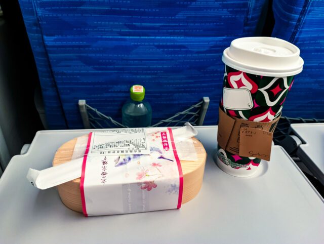 A wrapped ekiben bento box with chopsticks, a takeaway coffee cup, and a bottled drink on the fold-out tray of a Shinkansen seat with blue upholstery.