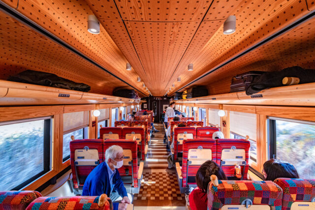 Interior of the Fujisan View Express train car 2 featuring wooden panels, colorful patterned seats, and passengers enjoying the ride.