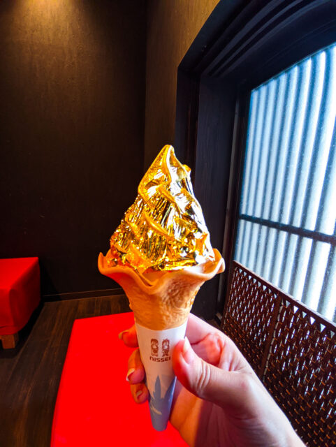 A hand holding a soft-serve ice cream cone wrapped in shimmering edible gold leaf from Hakuichi Higashiyama in Kanazawa, against a dark interior with red bench and shoji-style window.