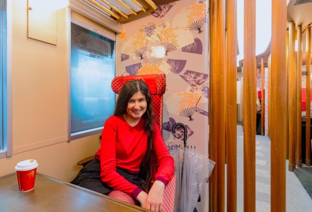 A woman in a red sweater smiles while seated inside the Hanayome Noren sightseeing train in Ishikawa, Japan. The carriage is decorated with elegant fan-patterned wallpaper, warm wooden lattice partitions, and distinctive red-and-black polka dot seats, reflecting traditional Kanazawa design aesthetics. A paper coffee cup and clear umbrellas rest nearby, adding to the cozy travel atmosphere.