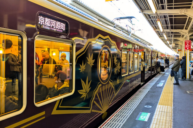 Exterior of the Hankyu Kyo Train at the station, featuring deep maroon body with golden fan and maple motifs, heading from Osaka Umeda to Kyoto-Kawaramachi.