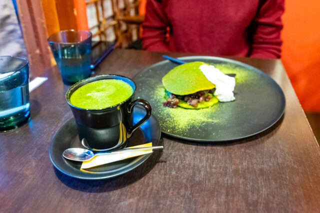 A black cup of frothy matcha latte on a saucer with a spoon, served beside a plate of matcha tiramisu layered with whipped cream and red beans, dusted generously with green tea powder, at Hayuwa café in Kanazawa’s Higashi Chaya District.