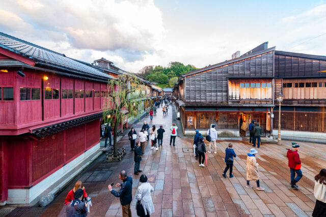 View from Hayuwa teahouse in Kanazawa, Japan, overlooking the historic Higashi Chaya District with its traditional wooden lattice-fronted buildings, red-painted teahouses, and stone-paved street filled with strolling visitors under the evening sky.