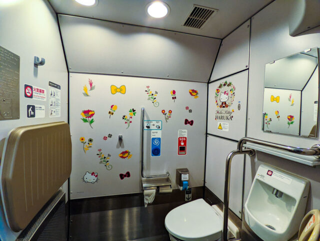 A restroom inside the Hello Kitty Haruka train, decorated with colorful floral stickers and Hello Kitty motifs, including bows and a cheerful Hello Kitty graphic on the wall. The toilet area is clean, modern, and bright, with both Western and urinal facilities.