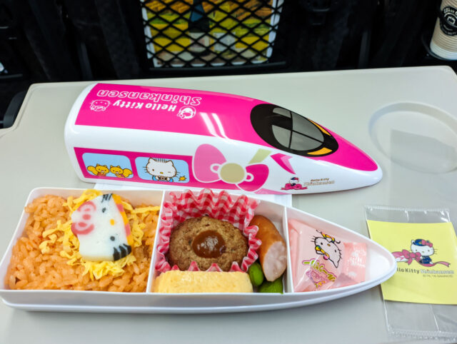 A colorful Hello Kitty Shinkansen bento box with rice, hamburger steak, egg, sausage, and Hello Kitty–themed packaging, served inside a train-shaped container.