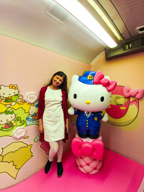 A woman with long dark hair, in creamy dress posing next to a large Hello Kitty conductor statue inside the Hello Kitty Shinkansen’s pink-themed photo spot.