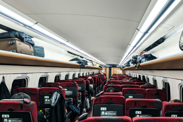 Interior of a Shinkansen bullet train in Japan with spacious seats, luggage racks, and passengers seated comfortably.