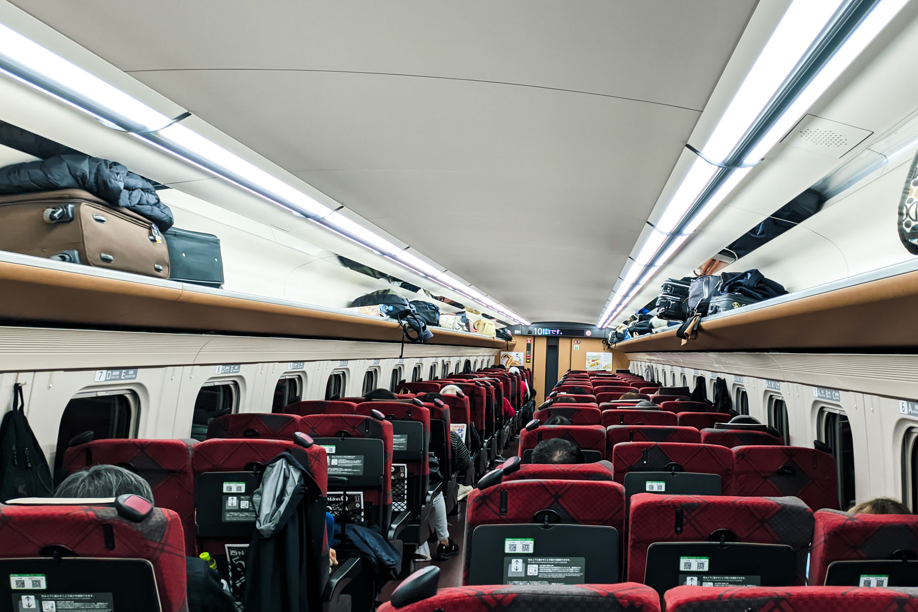 Interior of a Shinkansen bullet train in Japan with spacious seats, luggage racks, and passengers seated comfortably.