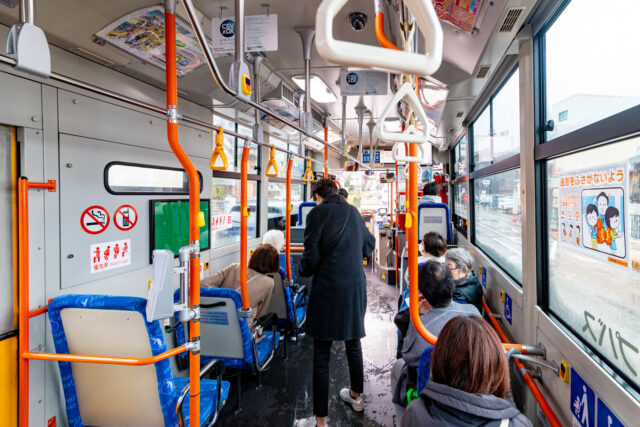 Inside view of a Japanese city bus with passengers seated, blue patterned seats, and bright orange handrails under natural daylight.