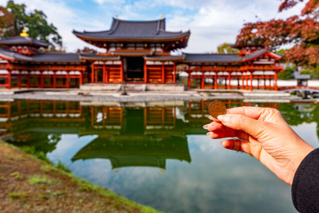 Hand holding a 10 yen Japanese coin in front of Byodoin Temple in Uji, with the temple’s reflection visible in the pond.