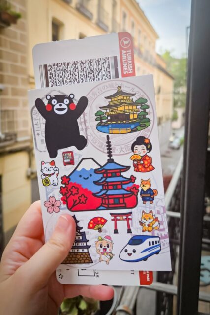 Hand holding a kawaii Japan-themed stampbook with cute stickers of Kumamon, Mount Fuji, a pagoda, geisha, shinkansen, and Japanese icons, with a boarding pass behind it.