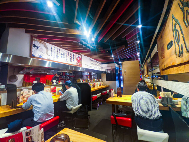 Inside a Japanese ramen shop Ippudo in Fukuoka with wooden counters, red accents, and customers seated while chefs prepare food behind the counter.