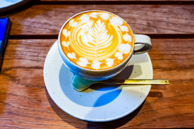 A cappuccino with detailed foam art in a white cup and saucer, served with a gold spoon at a café in Kumamoto.