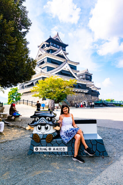 Smiling woman in a white and blue dress sitting next to a cute Kumamoto Castle mascot statue, with the iconic black-and-white Kumamoto Castle towering in the background on a sunny day in Japan.