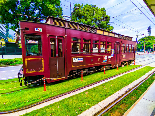 Retro-style maroon tramcar with gold detailing on green grass-lined tracks in Kumamoto, Japan, with trees and city buildings in the background.