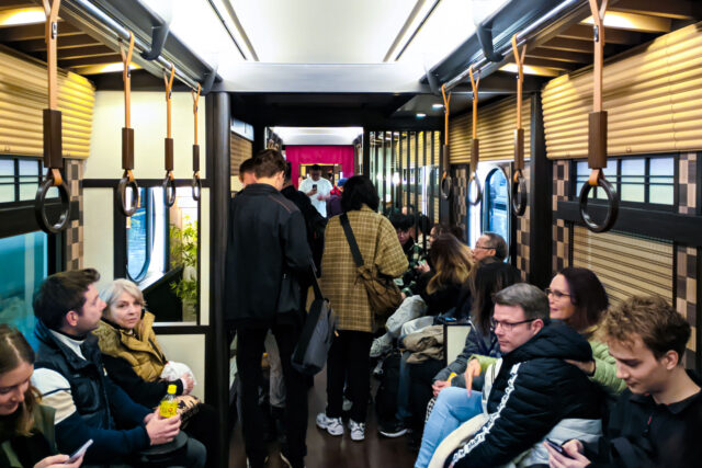 Crowded Interior of the Kyo Garaku train with passengers seated along wooden-style benches and standing, decorated in traditional Kyoto-inspired design with large windows.