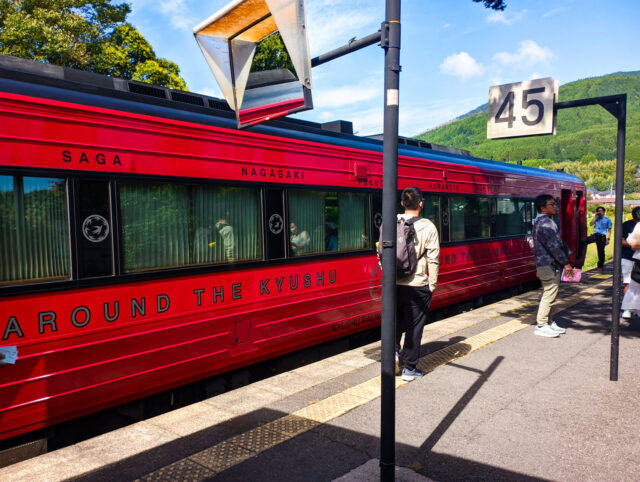 Bright red Yufu train of Kyushu railway stopped at a rural station platform, with passengers waiting nearby and green forested hills in the background.