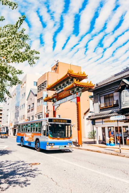 A city bus drives past the ornate red and gold gate of Nagasaki Chinatown, with a bright blue sky streaked with white clouds above and traditional-style buildings lining the street.