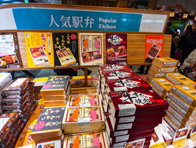 Display of popular ekiben (Japanese train station bento boxes) with various boxed meals stacked and labeled, ready for travelers.