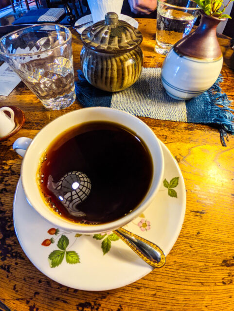 A cup of black coffee in a floral porcelain cup at Ranka Café in Takayama, with rustic wooden table, glass of water, and ceramic sugar p