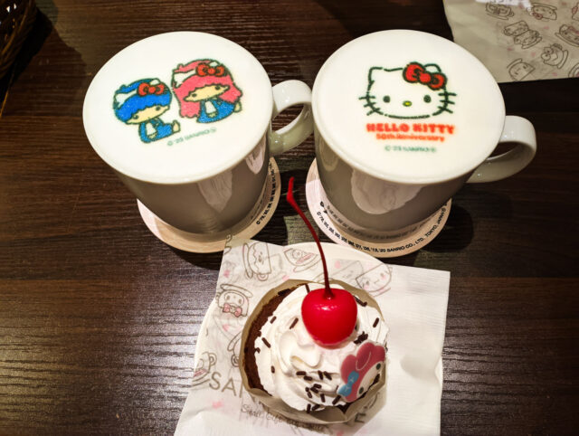 Character-themed drinks at Sanrio Café Ikebukuro, featuring cappuccinos with Hello Kitty and Little Twin Stars foam art, served alongside a small dessert - cupcake - with whipped cream and a cherry.