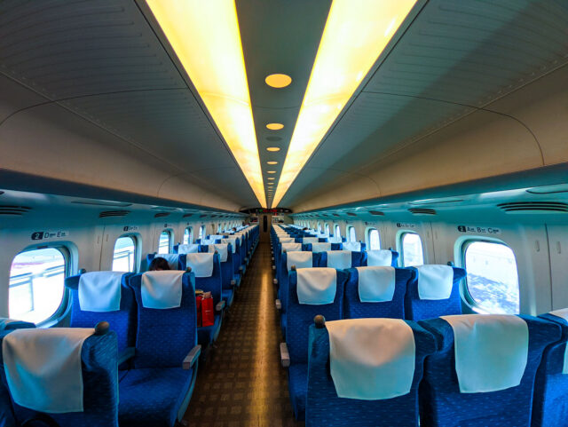Wide-angle view of a Shinkansen bullet train interior with rows of blue seats and white headrest covers, large windows on both sides, and bright overhead lighting.