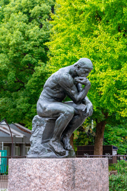 Bronze replica statue of The Thinker by Auguste Rodin on a stone pedestal, surrounded by green trees in Ueno Park’s museum area.