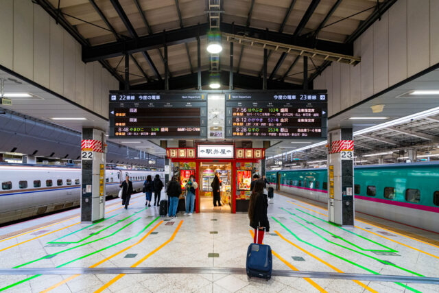 Passengers with luggage wait on the Shinkansen platform at Tokyo Station, standing between tracks 22 and 23 with bullet trains on both sides and an ekiben shop in the center.