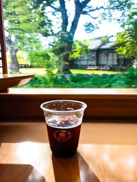 A cup of iced coffee on the wooden counter of the Two Stars 4047 sightseeing train in Kyushu, with sunlight shining through the window and a view of trees and a traditional wooden house passing by outside.