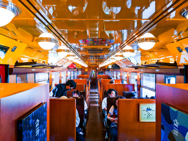 Passengers seated in the stylish wooden interior of the JR Kyushu Two Stars 4047 sightseeing train. The carriage features glossy yellow ceilings, warm wood partitions, and soft globe lights, creating a cozy retro atmosphere.