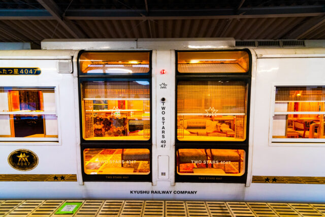 The luxurious Two Stars 4047 sightseeing train by Kyushu Railway Company, showing its elegant white exterior and large windows that reveal the warm, golden-lit lounge-style interior.