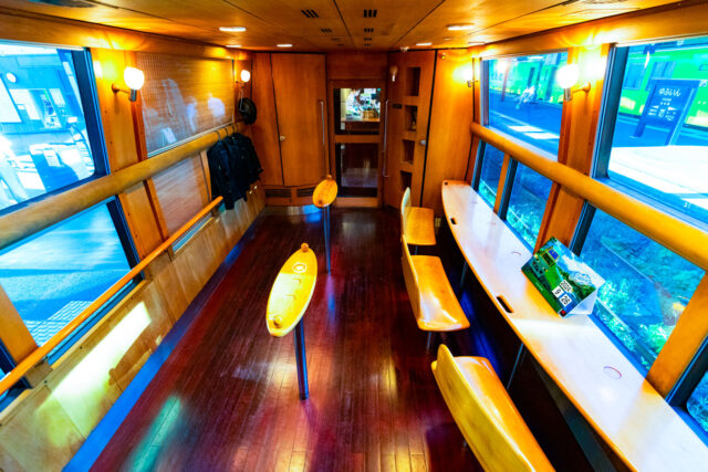 Inside the Yufuin no Mori sightseeing train in Kyushu, Japan, showing its warm wooden interior with counter seating by large panoramic windows and small standing tables, designed for passengers to relax and enjoy the scenic views along the route.