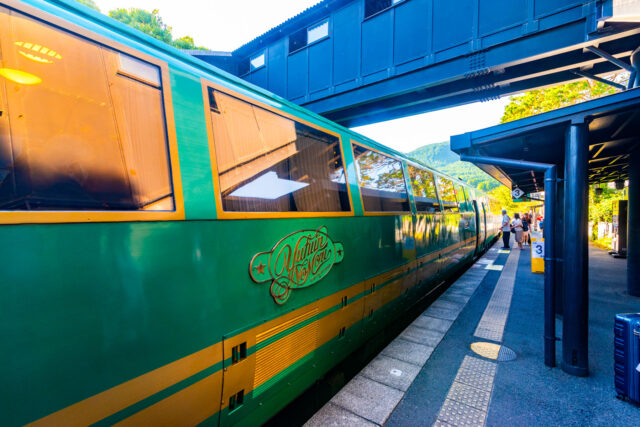The Yufuin no Mori sightseeing train in Yufuin Kyushu, Japan, with its signature green and gold exterior reflecting the sunlight, waiting at a rural station platform against a backdrop of lush mountains.